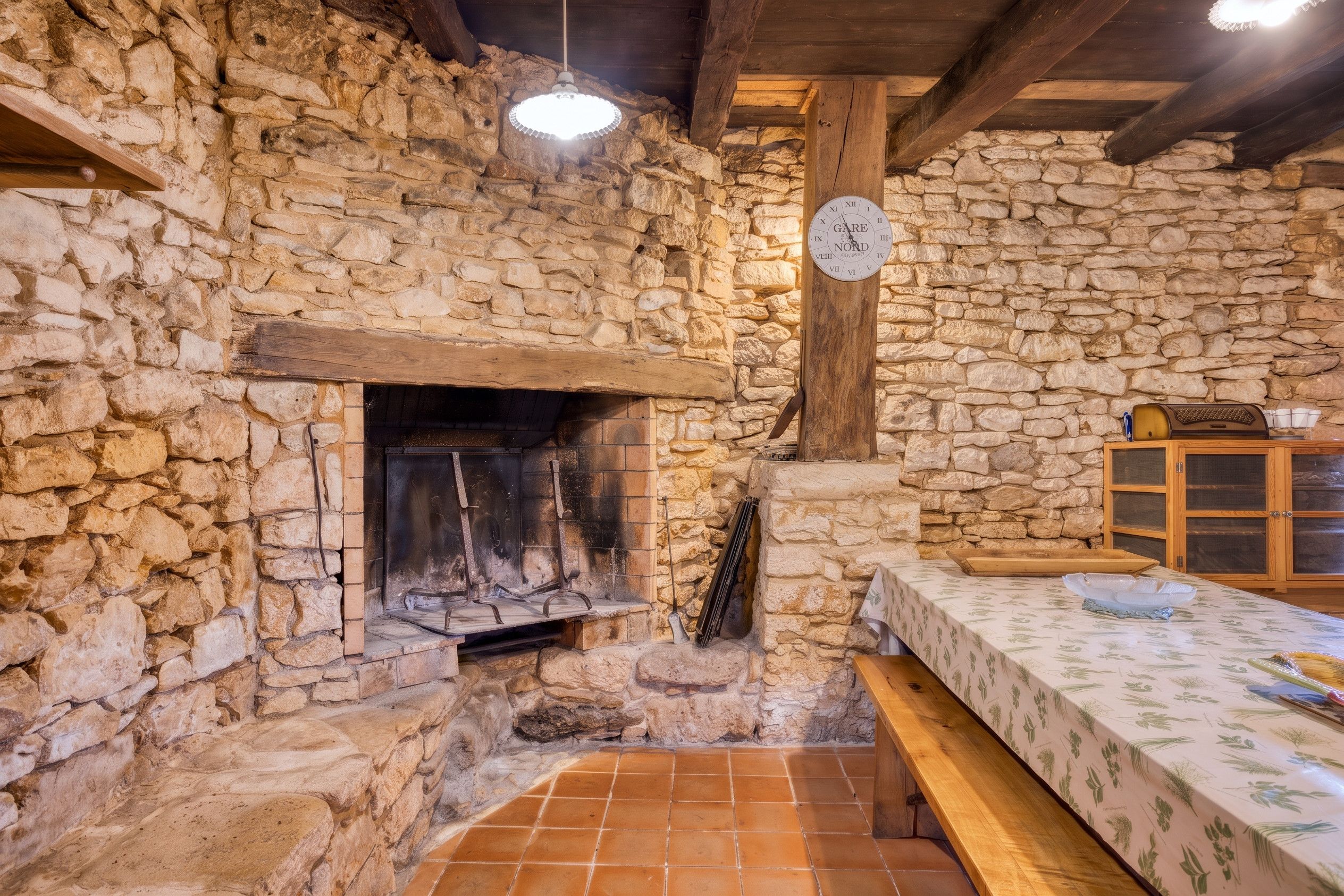 Une pièce aux murs de pierre, avec une cheminée, un banc en bois, une table recouverte d’une nappe à motifs floraux et une horloge murale.