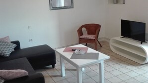 A living room with a black sofa, a white coffee table, a red chair, a framed picture on the wall, a TV on a white stand, and a tiled floor.