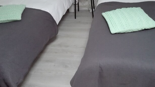 A room with two beds, each with a grey bedspread and a light green throw. There is a small black stool between the beds.