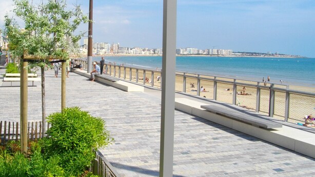 A beachside promenade with a wooden deck, seating areas, and a view of the beach and cityscape.