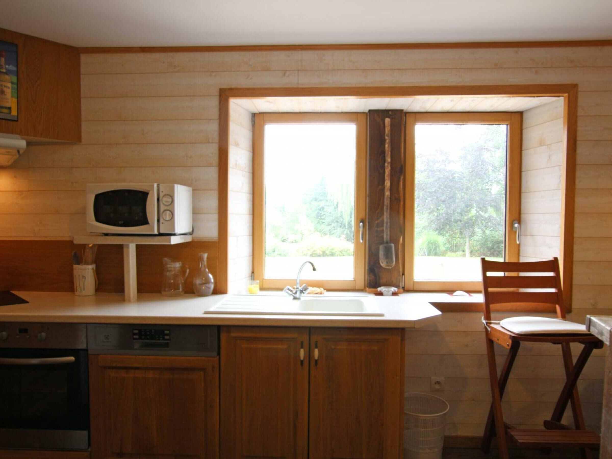 Eine kleine Küche mit Holzschränken, einer Mikrowelle, einem Spülbecken und einem Fenster mit Blick auf Grünflächen.