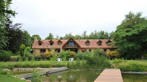 A large, rustic-style building with multiple gabled roofs and a wooden deck leading to a pond. - Charming house with garden and parking in Val-Saint-Père (Le Val-Saint-Père)