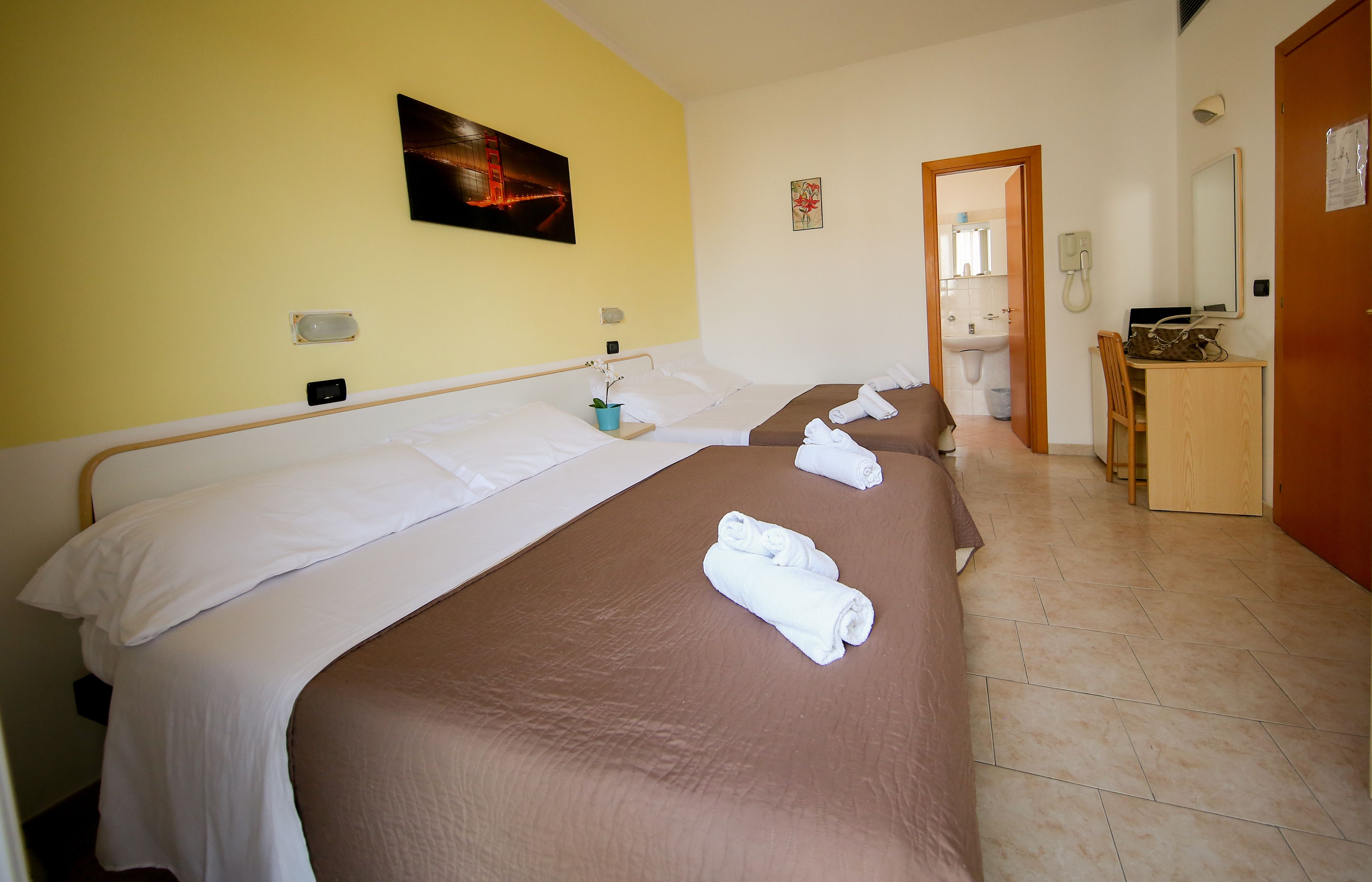A hotel room with two beds, a desk, a chair, a bathroom visible through an open door, and a painting on the wall.