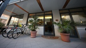Un edificio moderno con amplia entrada de vidrio, plantas en macetas y bicicletas estacionadas afuera.