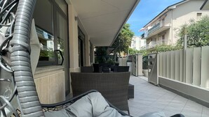 Un espacio de estar al aire libre cubierto, con una silla acolchada y una mesa, piso de baldosas y un edificio al fondo.