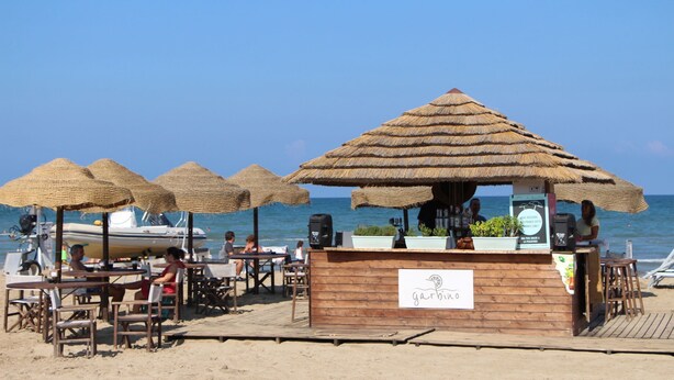 Un bar en la playa con sombrillas de paja y mesas de madera.