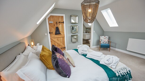 A bedroom with a bed, pillows, a hanging light fixture, a chair, and a door leading to a bathroom. - Wild Cherry Barn, Ashlin Farm Barns (Lincoln)