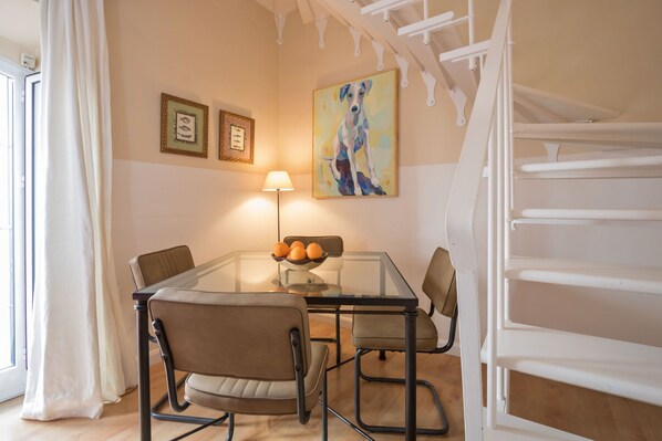 A dining area with a glass table, four chairs, a bowl of oranges, and a painting on the wall.