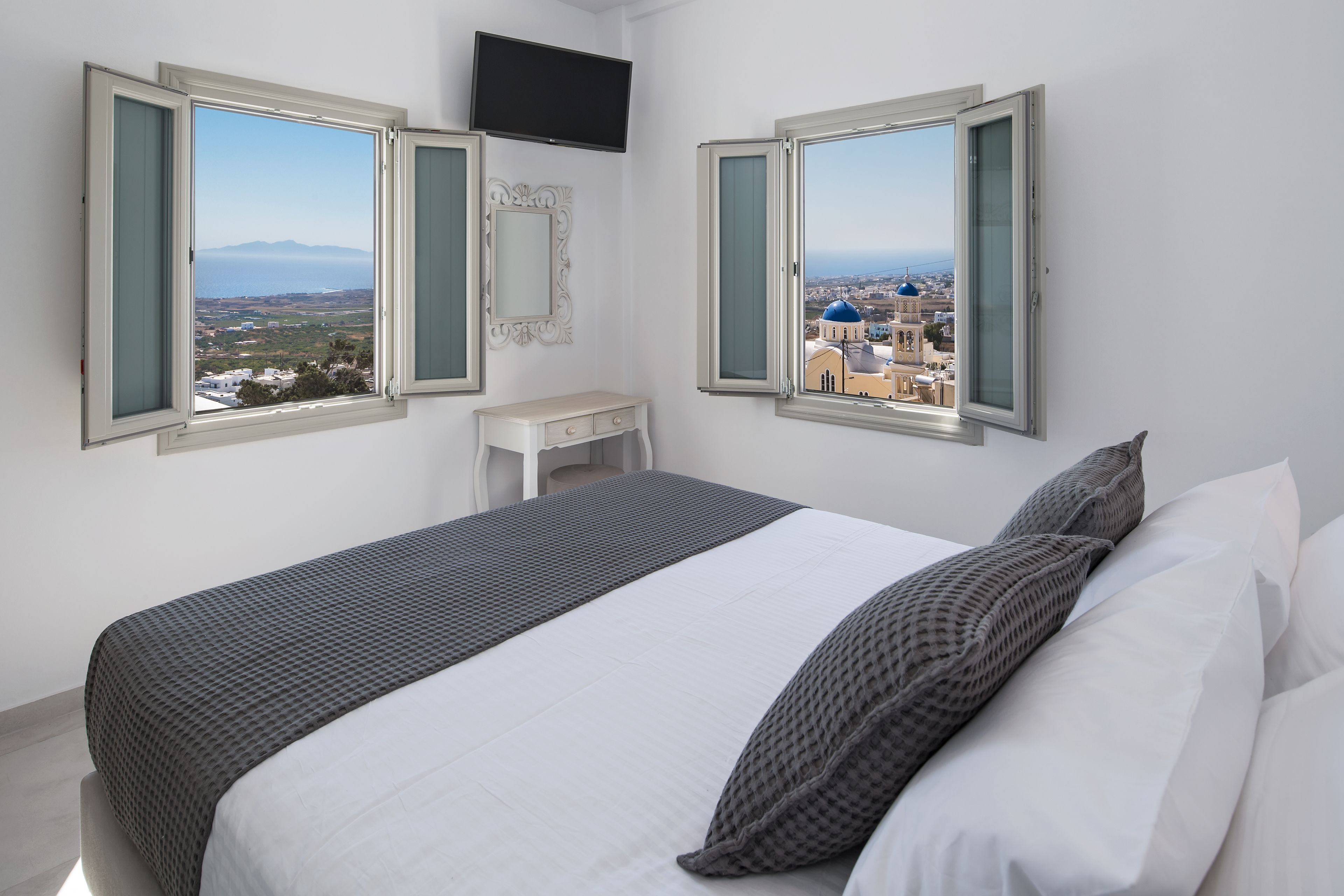 Una habitación de hotel con una cama, dos ventanas con contraventanas abiertas, una mesita de noche pequeña y un televisor montado en la pared.