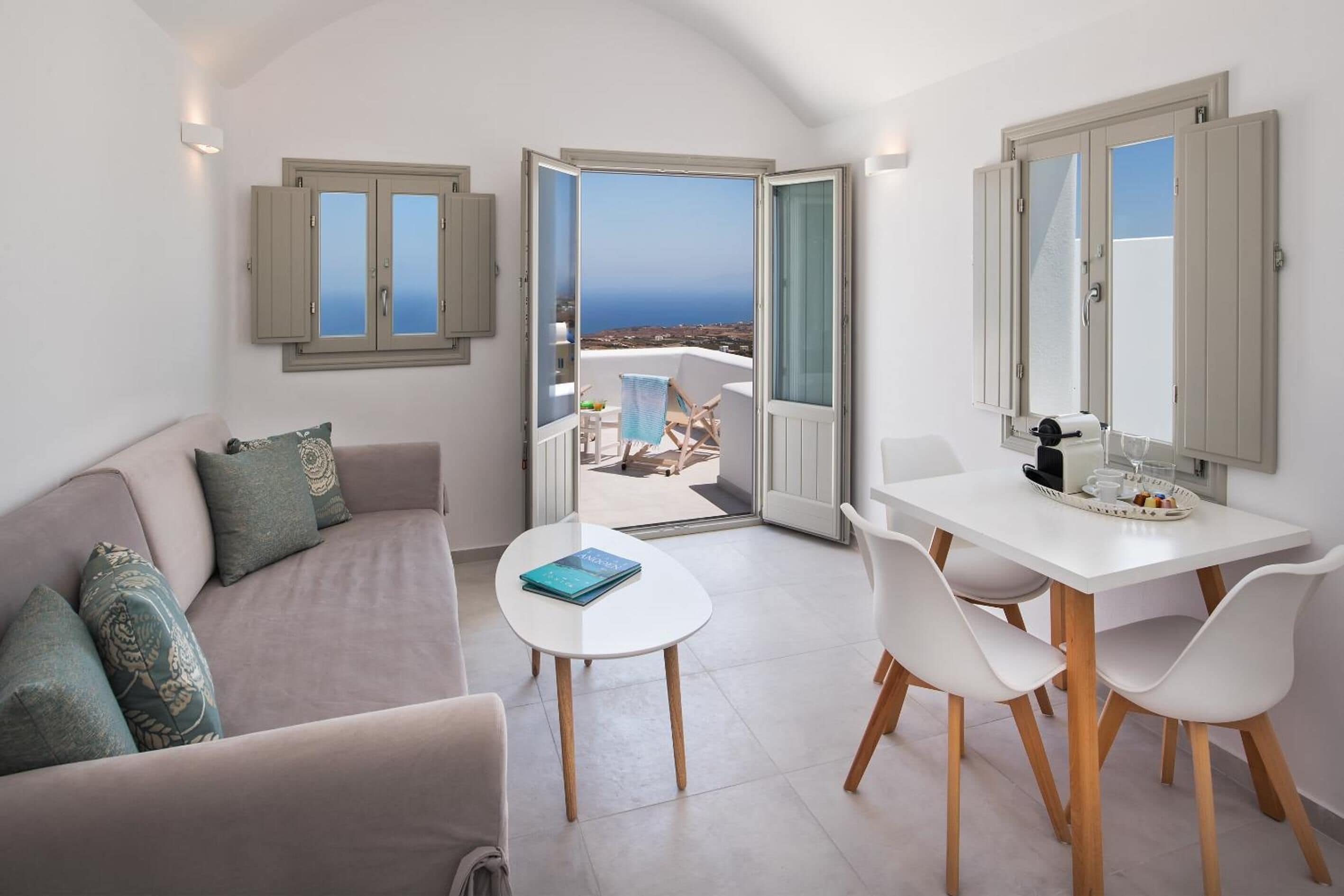 Een moderne woonkamer met een bank, een klein rond tafeltje en een eethoek met stoelen. Er zijn twee ramen met luiken, waarvan er één uitkomt op een balkon met uitzicht op zee.