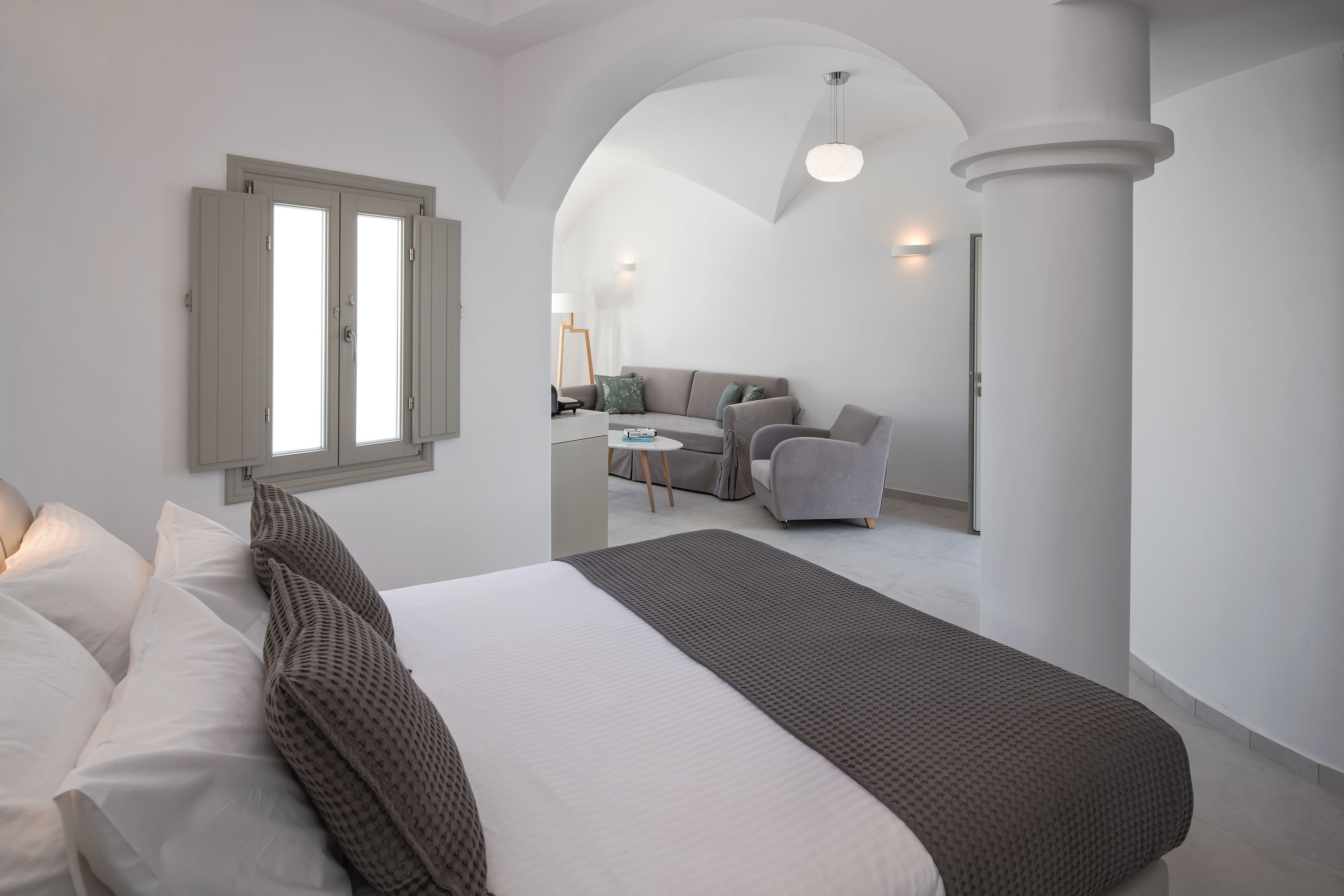 A modern hotel room with a large bed, a seating area with a sofa and chairs, and a view through an arched doorway to another room.