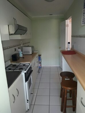 Une petite cuisine étroite équipée d’une cuisinière à gaz, d’un four à micro-ondes et d’un petit plan de travail. On y trouve deux tabourets et un meuble sur lequel est posé un four à micro-ondes.