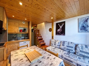 A cozy cabin interior with a wooden ceiling, a kitchenette, a sofa with patterned cushions, and a deer painting on the wall.