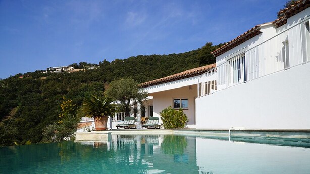 A poolside view of a white building with a tiled roof, surrounded by lush greenery and a clear blue sky.