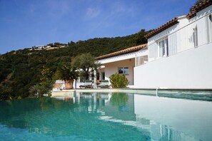 A poolside view of a white building with a tiled roof, surrounded by lush greenery and a clear blue sky.