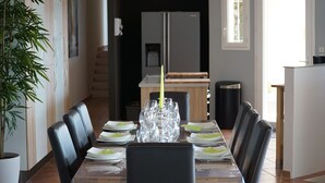A dining area with a long table set for six, surrounded by black chairs, with a potted plant to the left.