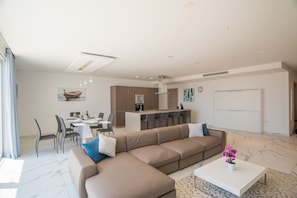 Uma sala de estar moderna com um sofá seccional bege grande, uma área de jantar com mesa e cadeiras brancas e uma cozinha com armários de madeira e bancada branca.