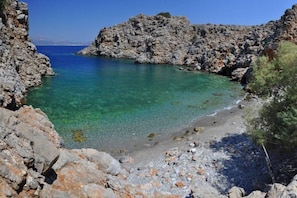 A rocky coastline with clear turquoise water, a pebble beach, and sparse vegetation.