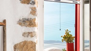 A room with a stone wall, a red-framed window, a wooden chair, and a table with a blue object on it.