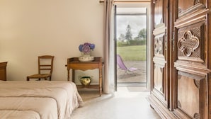 Um quarto com uma cama, uma cadeira de madeira, uma mesa pequena com um vaso de flores e vista para um jardim através de uma porta de vidro.