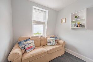 Um sofá bege com almofadas decorativas, uma janela com persianas, uma estante de livros e uma planta, e um tapete cinza.