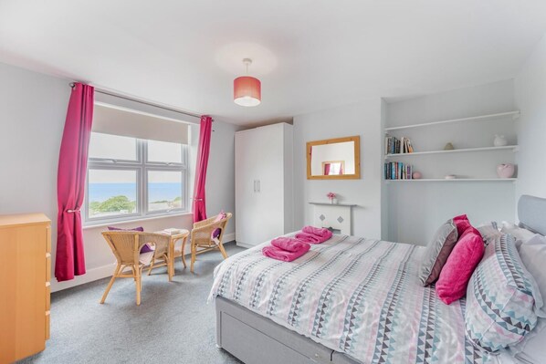 A bedroom with a bed, a desk, a chair, a wardrobe, a mirror, and a view of the sea.
