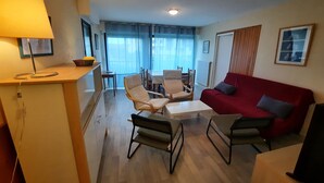 Una sala de estar moderna con un sofá rojo, dos sillones, una mesa de centro y una lámpara. Se observa un comedor con una mesa y sillas al fondo.