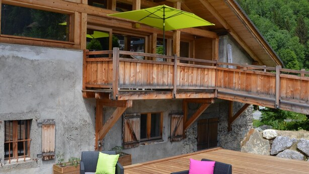 Un chalet en bois doté d’un balcon, d’un espace extérieur aménagé avec des meubles en osier, et offrant une vue sur une montagne boisée.