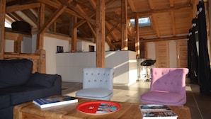 Un intérieur en bois avec un plafond haut, un canapé, un fauteuil de couleur claire et une table à manger avec des livres.