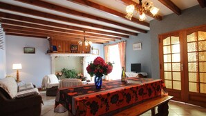 A cozy living room with a wooden bench, a table with a floral tablecloth, a vase with flowers, and a television on a stand.