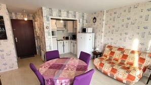 A compact living space with a dining table, purple chairs, a sofa, a kitchenette, and patterned wallpaper. - Charming apt with terrace, parking, near beach and shops (Les Sables-d'Olonne)