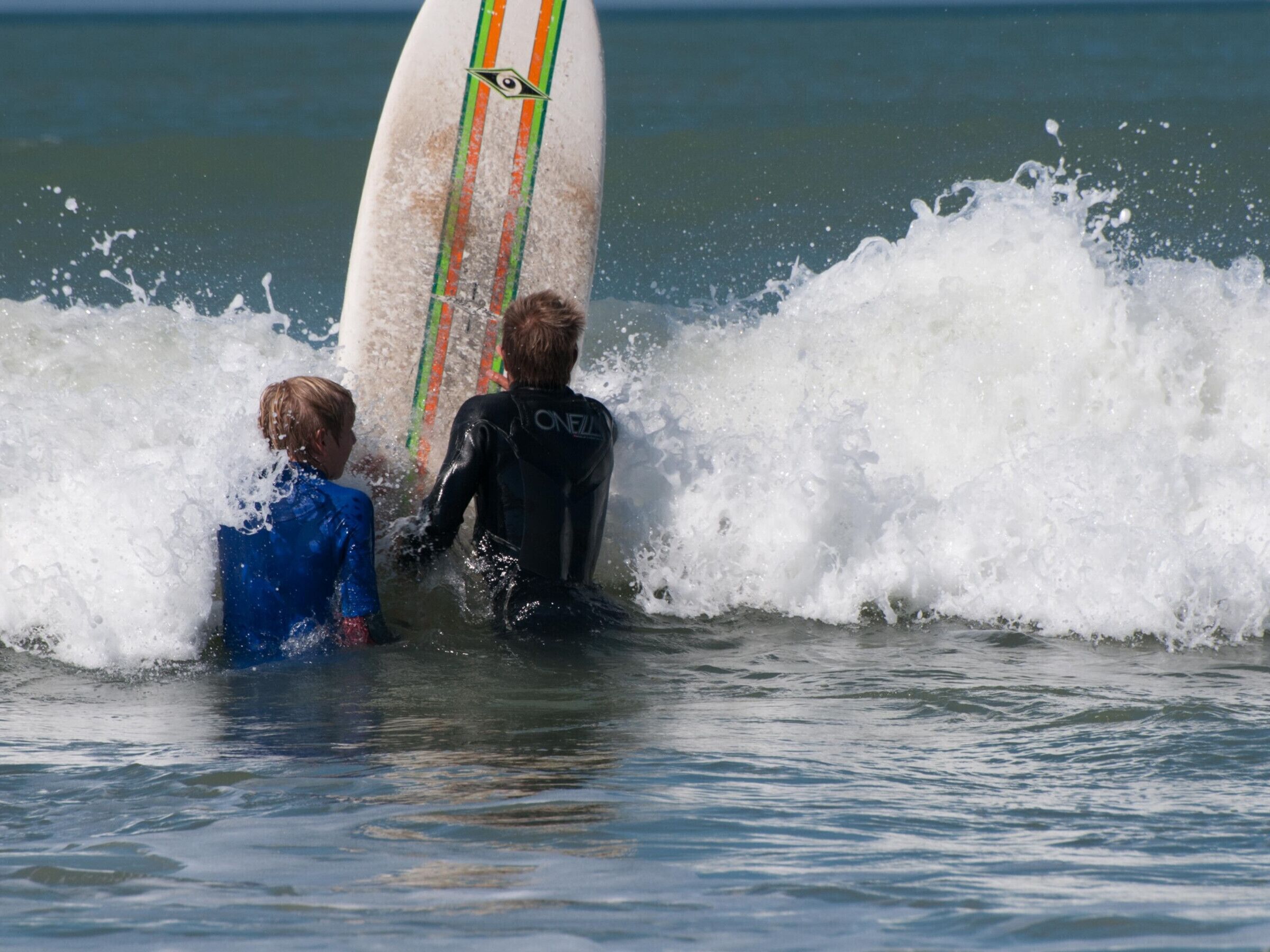 Dos personas con trajes de neopreno de pie en el agua con una tabla de surf.