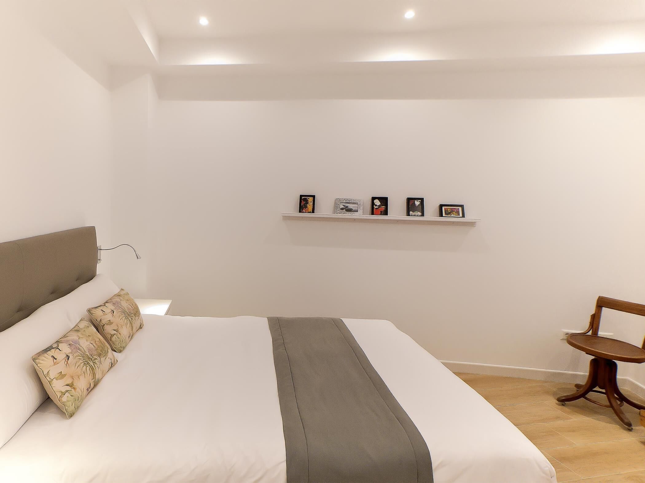 A bedroom with a bed, pillows, a bedside table, a chair, and a shelf with framed pictures.