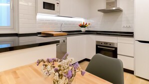 Een moderne keuken met witte kasten, een magnetron, fornuis en vaatwasser. Op het aanrecht ligt een houten snijplank en staat een vaas met bloemen.