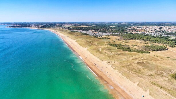 Eine Vogelperspektive einer Küstenregion mit einem Sandstrand, klarem türkisblauem Wasser und einer in der Ferne liegenden Stadtansicht.