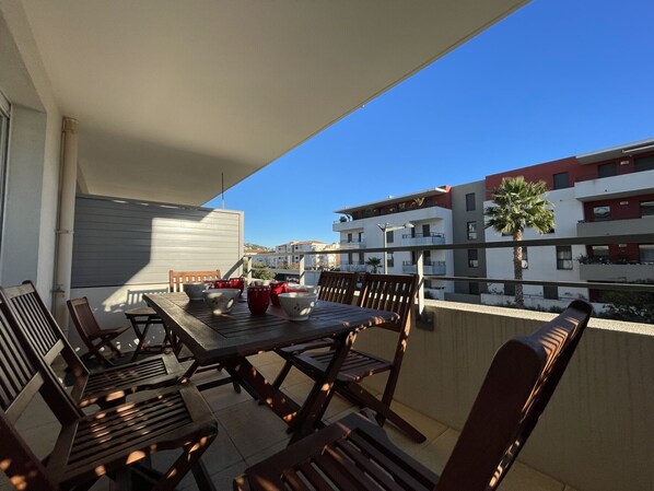 An outdoor balcony with a wooden table and chairs, a view of modern buildings, and a clear blue sky.