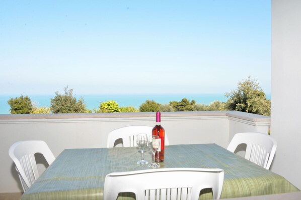A table with a bottle of red wine and two glasses, set against a backdrop of a clear blue sky and a distant view of the sea.