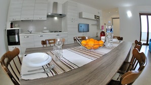 A modern kitchen with white cabinets, a built-in oven, and a large dining table set for six with striped placemats and a bowl of oranges.