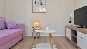 Un salon moderne avec un canapé rose, une petite table ronde surmontée d’une lampe, un tapis blanc et un téléviseur posé sur un meuble en bois.