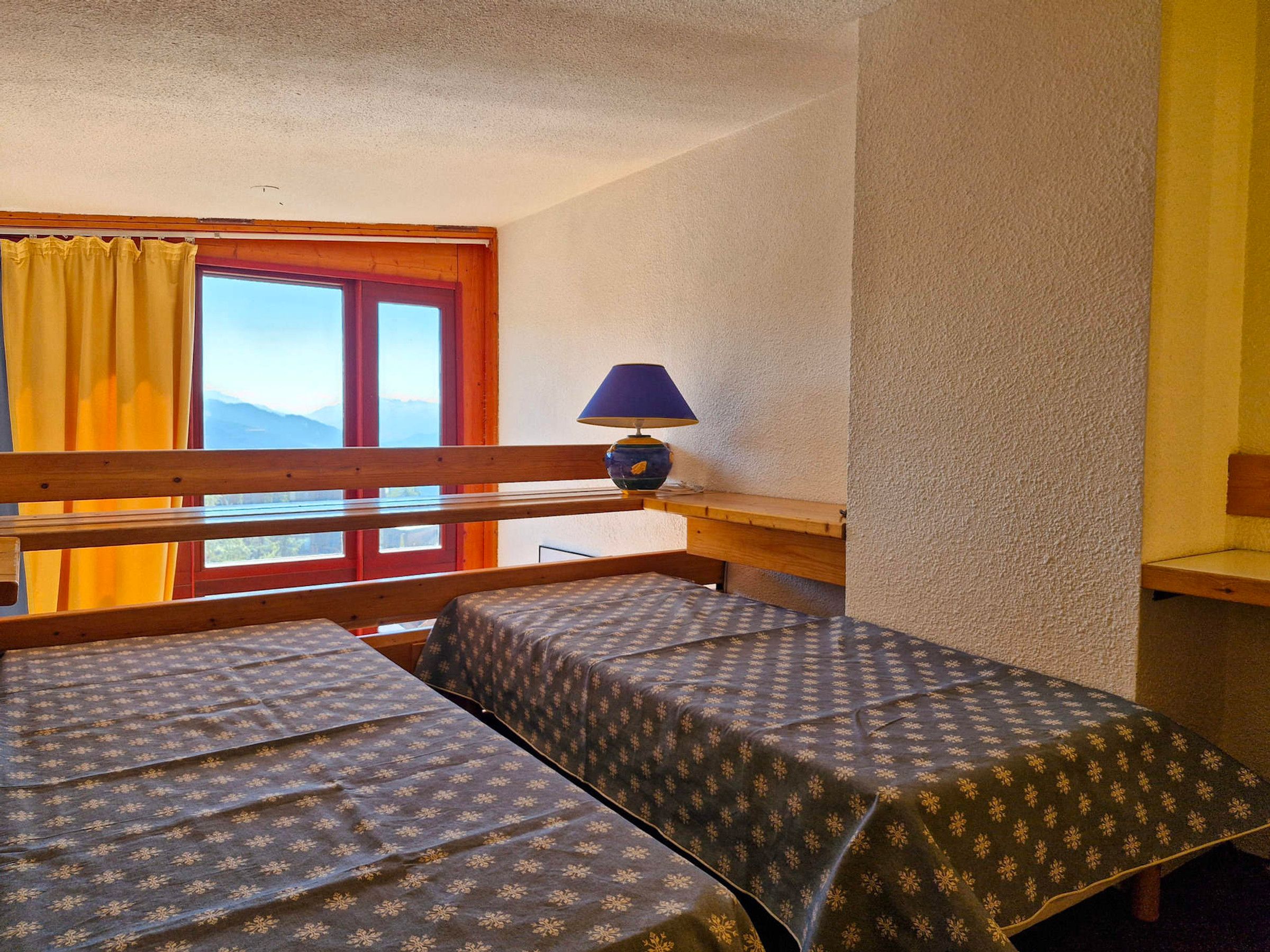 Quarto com duas camas, janela com cortinas, luminária e vista para montanhas.
