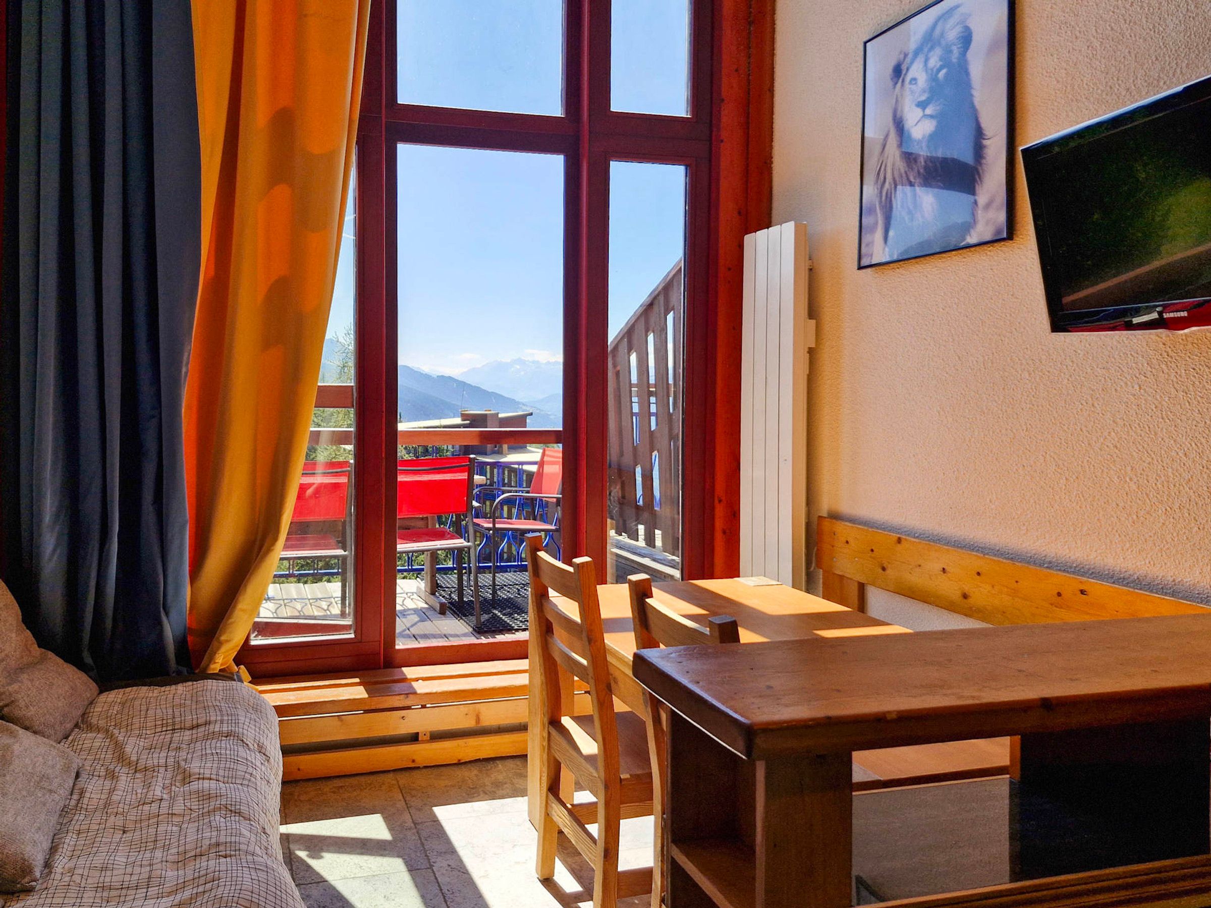 Ein Zimmer mit Holztisch und Stühlen, einem fenster mit Vorhängen, einem Gemälde an der Wand und Blick auf Gebäude und Berge.