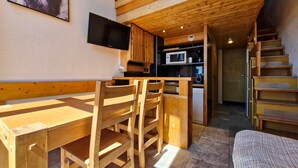Un interior de madera con una mesa de comedor, sillas, un televisor montado en la pared, una zona de cocina con electrodomésticos y una escalera.