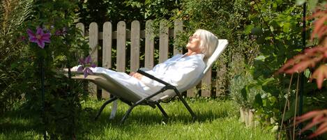 Eine Person lehnt in einem Gartenstuhl zurück, im Hintergrund ist ein Holenzaun und viel Grünwerk zu sehen.