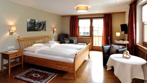 Ein Hotelzimmer mit einem hölzernen Bett, zwei Nachttischen, einem Schreibtisch, einem Sessel, einem kleinen Tisch mit Blumenvase und einem Bild an der Wand.