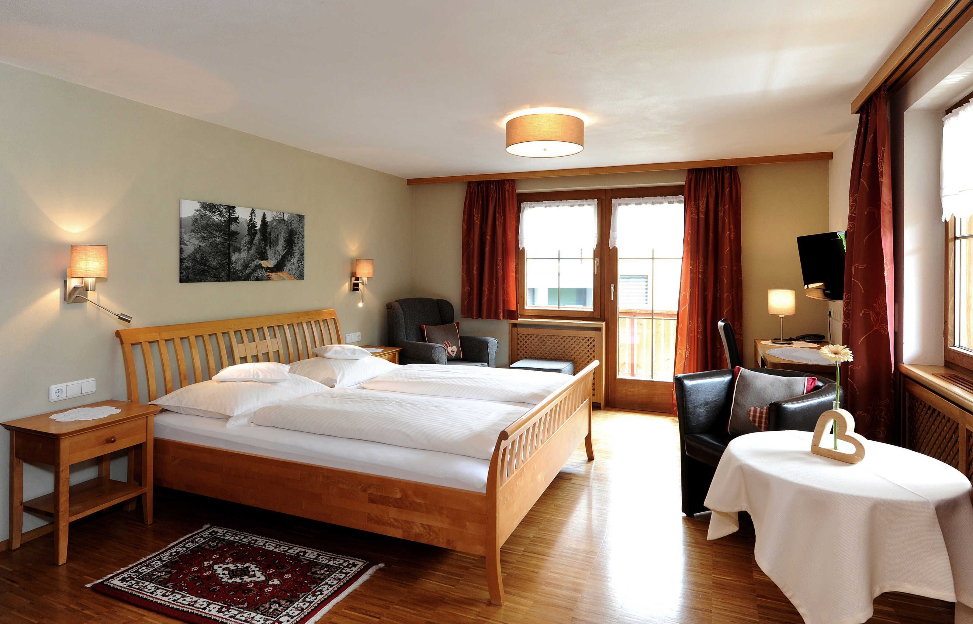 A hotel room with a wooden bed, two bedside tables, a desk, a chair, a small table with a flower vase, and a painting on the wall.