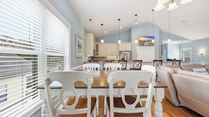 A modern kitchen with white cabinets, a dining area with white chairs, and a living room with a beige sofa.