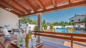 Une table en bois sur laquelle se trouvent un plateau avec des verres et des bouteilles, un télescope et un banc. Au fond, on aperçoit une piscine et une maison avec un toit rouge.