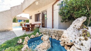 Un espace piscine agrémenté d’une fontaine en pierre, une table à manger avec des chaises, et une vue sur une maison avec un toit bleu.