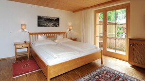 Un dormitorio con una cama de madera, dos mesitas de noche, una mesita auxiliar, una alfombra roja, un ventanal con vistas y un cuadro colgado en la pared.