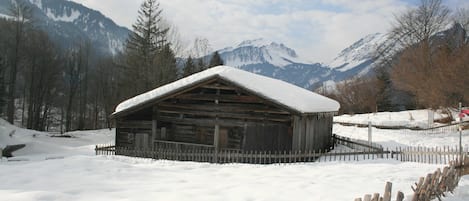 A wooden cabin with a snow-covered roof, surrounded by a wooden fence, set against a backdrop of snow-covered mountains and trees.